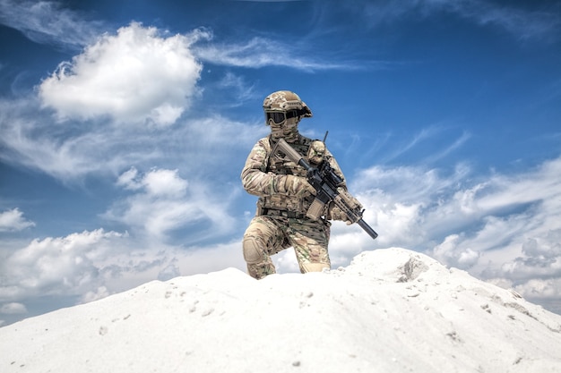 Hombre con uniforme de camuflaje militar con réplica de rifle de servicio, de pie sobre una duna de arena con cielo nublado en el fondo, imitando al tirador de las fuerzas especiales del ejército de EE. UU. Durante los juegos de guerra de airsoft en el desierto