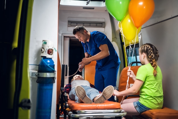 Un hombre con uniforme médico le pone una máscara de oxígeno a una niña acostada en una camilla, su hermana pequeña está mirando al médico trabajar.