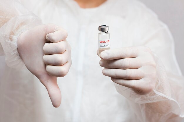 Un hombre con uniforme médico sostiene una ampolla con una vacuna contra el coronavirus frente a él y da un pulgar hacia abajo. Contra la vacunación frente al covid-19.