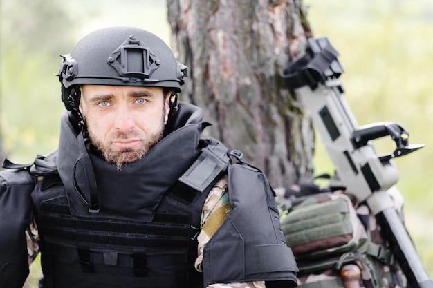 Un hombre con uniforme militar y un chaleco antibalas se sienta en el  bosque cerca de un detector de metales y una mochila militar un hombre hace  una pausa en el trabajo