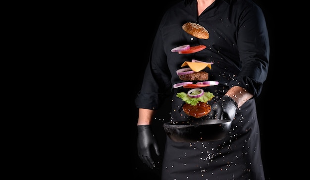 Hombre con uniforme negro sosteniendo una sartén redonda de hierro fundido con ingredientes levitando hamburguesa con queso