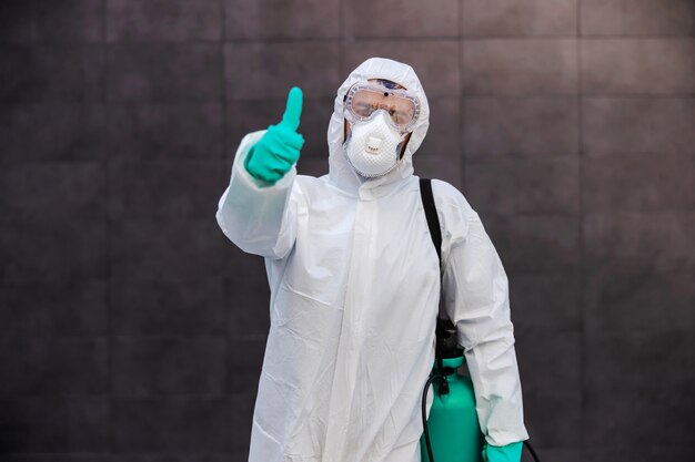 Hombre en uniforme protector estéril con guantes de goma que llevan rociador con desinfectante y mostrando los pulgares hacia arriba mientras está de pie al aire libre. Prevención de la propagación del concepto de virus corona.