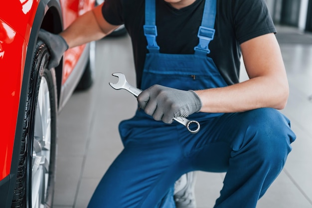El hombre en uniforme se sienta con una llave en la mano cerca de un automóvil roto Concepción del servicio de automóviles