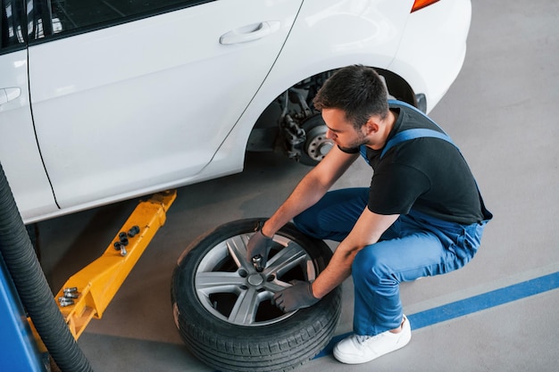 Hombre con uniforme de trabajo sentado y cambiando la rueda del coche en el interior Concepción del servicio de automóviles