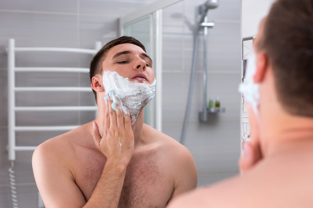 Hombre untando espuma para afeitarse en la cara de pie delante del espejo en el moderno baño de azulejos en casa