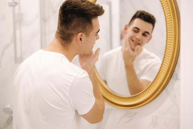 El hombre usa espuma de afeitar en el baño. Un hombre guapo con camiseta blanca se afeita por primera vez.