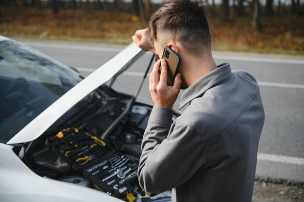 El hombre usa un garaje de llamadas de teléfono celular frente al capó abierto de un automóvil roto en la carretera en el bosque Concepto de avería del automóvil