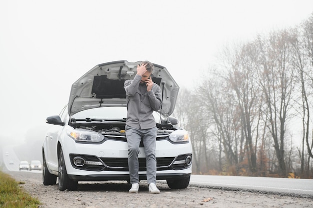 El hombre usa un garaje de llamadas de teléfono celular frente al capó abierto de un automóvil roto en la carretera en el bosque Concepto de avería del automóvil