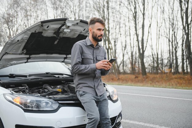 El hombre usa un garaje de llamadas de teléfono celular frente al capó abierto de un automóvil roto en la carretera en el bosque Concepto de avería del automóvil