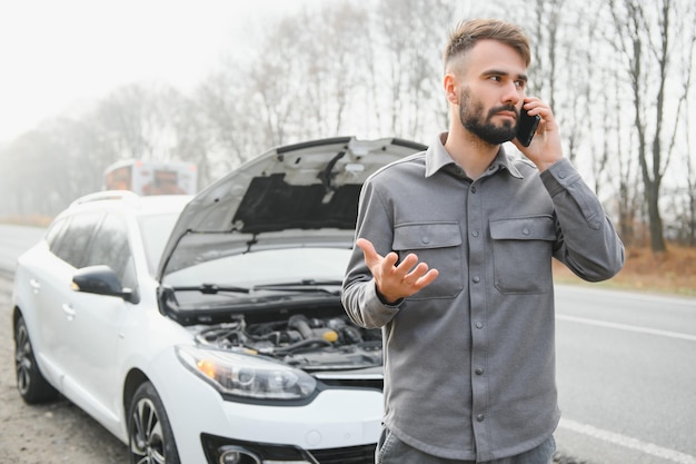 El hombre usa un garaje de llamadas de teléfono celular frente al capó abierto de un automóvil roto en la carretera en el bosque Concepto de avería del automóvil