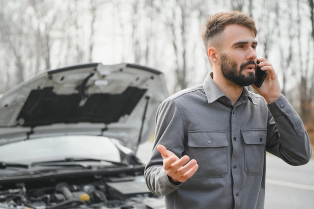 El hombre usa un garaje de llamadas de teléfono celular frente al capó abierto de un automóvil roto en la carretera en el bosque Concepto de avería del automóvil