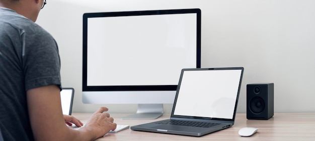Hombre usando una computadora de escritorio y una computadora portátil en la oficina en casa maqueta pantalla blanca en blanco