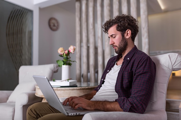 Hombre usando la computadora portátil, trabajando en casa. Preocupado por las finanzas. Hombre guapo con barba trabajando en casa en proyecto, está sentado en el sofá mirando su computadora portátil frente a él. Centrarse en el hombre