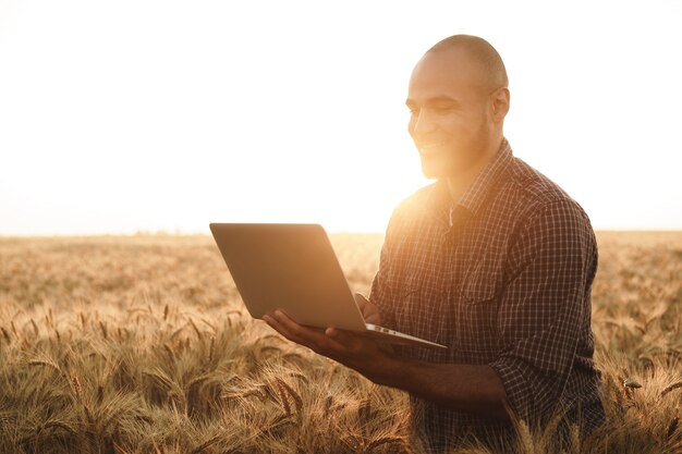 Hombre usando laptop mientras está de pie en el campo de trigo al atardecer