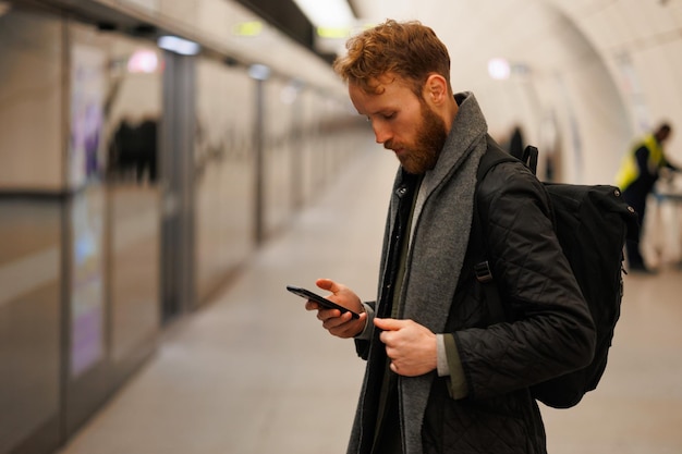 Hombre usando un teléfono inteligente en la plataforma de la estación de metro mientras espera el tren