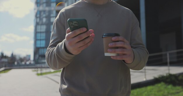 Hombre usando un teléfono inteligente y sosteniendo café para llevar caminando por la calle de la ciudad con edificios en el fondo Concepto de personas y comunicación