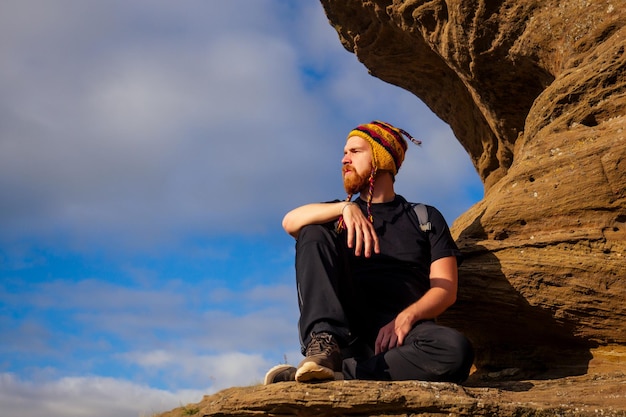 Hombre valiente pelirrojo barba escalada búlder turista de roca de piedra sube con mochila en una camiseta negra y un sombrero divertido hecho de lana de yak de fondo de cielo de Nepal