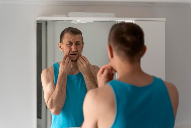 El hombre se ve descontento con su reflejo en el espejo por la mañana en el baño Cuidado de la piel facial y afeitado