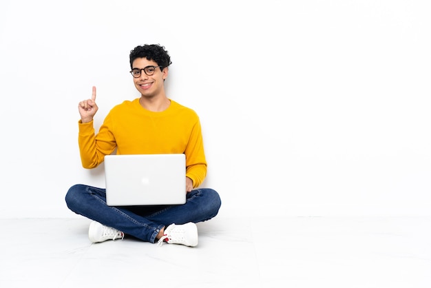 Hombre venezolano sentado en el suelo con laptop mostrando y levantando un dedo en señal de lo mejor