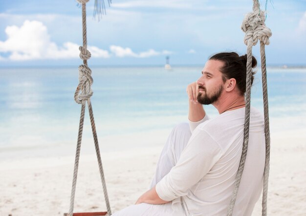 Hombre vestido de blanco sentado en el columpio de la playa y soñando