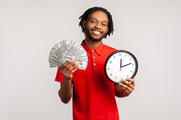 Hombre vestido con camiseta roja de estilo casual que muestra un dólar en efectivo y un reloj de pared en la cámara pago por hora