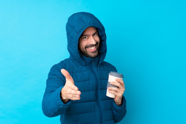 Hombre vestido con chaqueta de invierno y sosteniendo un café para llevar sobre una pared azul aislada estrechándole la mano para cerrar un buen trato