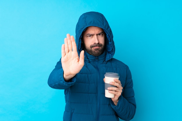 Hombre vestido con chaqueta de invierno y sosteniendo un café para llevar sobre la pared azul aislada haciendo gesto de parada