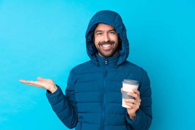 Hombre vestido con chaqueta de invierno y sosteniendo un café para llevar sobre la pared azul aislado con copyspace imaginario en la palma para insertar un anuncio