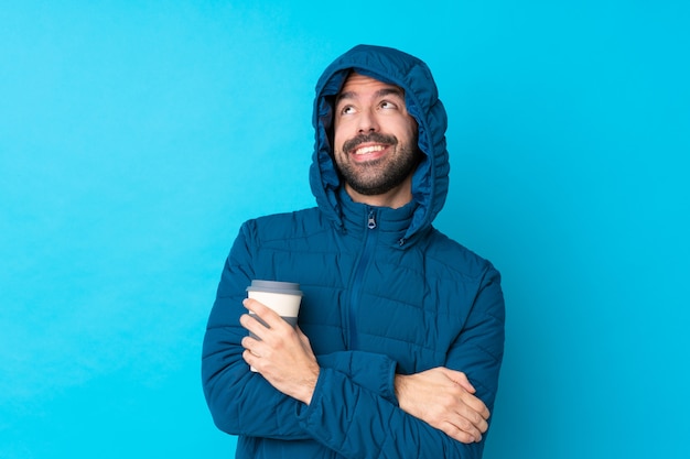 Hombre vestido con chaqueta de invierno y sosteniendo un café para llevar sobre la pared azul aislado mirando hacia arriba mientras sonríe