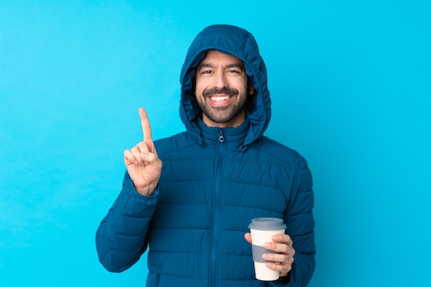 Hombre vestido con chaqueta de invierno y sosteniendo un café para llevar sobre pared azul aislado mostrando y levantando un dedo en señal de lo mejor