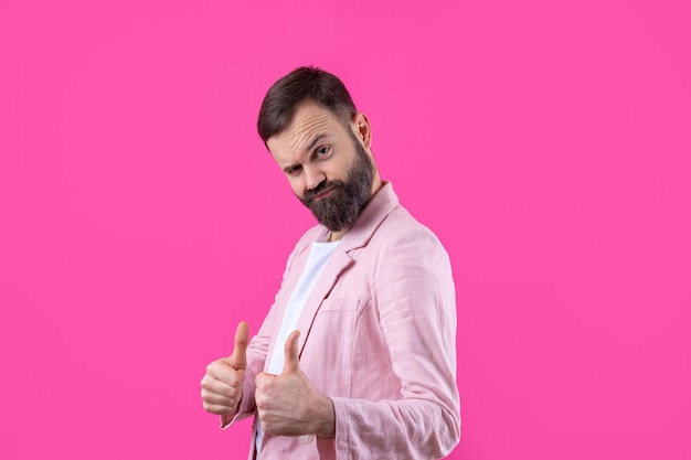 Un hombre vestido con una chaqueta rosa con barba muestra un pulgar hacia arriba
