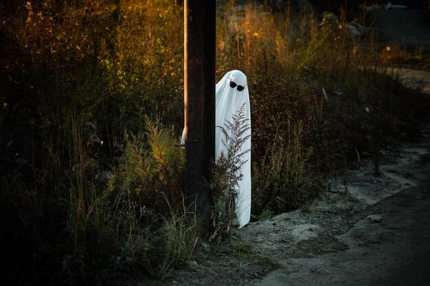 Un hombre vestido como un fantasma de una sábana blanca se esconde detrás de un poste junto a la carretera Ghost Challend 2021 Celebración de Halloween Temporada espeluznante