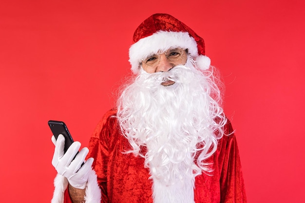 Hombre vestido como Santa Claus consultando su teléfono móvil sobre fondo rojo Celebración de Navidad regalos concepto de consumismo y felicidad