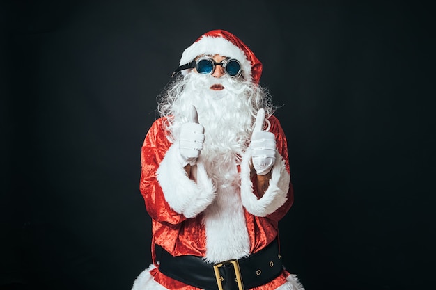 Hombre vestido como Santa Claus con gafas de soldador de estilo victoriano, levantando los pulgares hacia arriba, sobre fondo negro. Concepto de Navidad, Santa Claus, regalos, celebración.