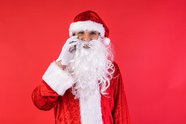 Hombre vestido como Santa Claus hablando por su teléfono móvil sobre fondo rojo Celebración navideña regalos concepto de consumismo y felicidad