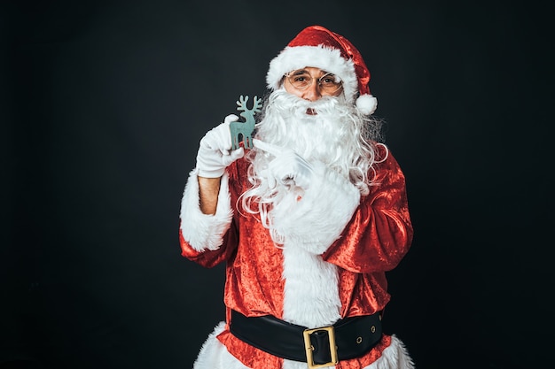 Hombre vestido como Santa Claus sosteniendo la figura de un reno de madera verde, sobre fondo negro. Concepto de Navidad, Santa Claus, regalos, celebración.