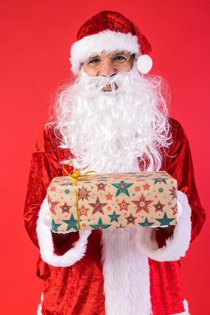 Hombre vestido como Santa Claus sosteniendo un regalo sobre fondo rojo Celebración navideña regalos concepto de consumismo y felicidad