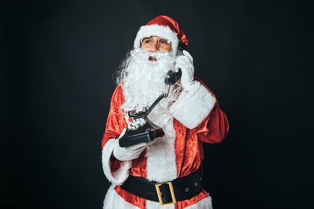 Hombre vestido como Santa Claus sosteniendo un teléfono retro de los años 60, hablando, sobre fondo negro. Concepto de Navidad, Santa Claus, regalos, celebración.