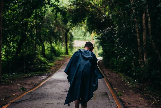 Hombre vestido con un impermeable caminando en medio de un camino al lado del bosque