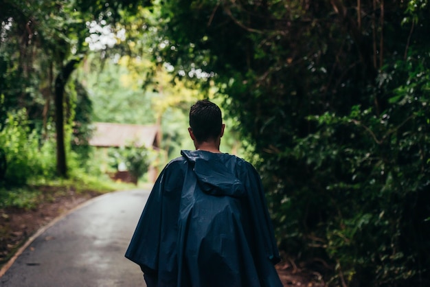 Hombre vestido con un impermeable caminando en medio de un camino al lado del bosque