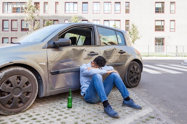 Un hombre vestido con jeans y camisa azul sentado cerca de un auto chocado con una botella de bebida alcohólica y durmiendo en un automóvil dañado, una infracción por conducir en estado de ebriedad provocó un accidente de tráfico. Disparo al aire libre.