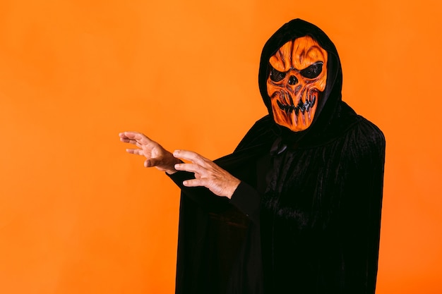 Foto hombre vestido con máscara de látex de calabaza y capa de terciopelo con capucha, asusta con las manos, sobre fondo naranja. halloween y días de los muertos concepto.