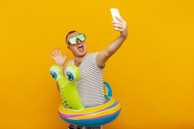 Hombre vestido con máscara submarina camisa a rayas nadar vueltas mirando al teléfono tomando selfie muy emocionalmente mientras sonríe en concepto de vacaciones de espacio amarillo