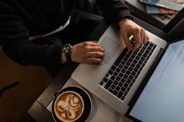 Hombre vestido de negro con un reloj de lujo trabaja en una computadora portátil y toma café en una cafetería. Chico con computadora y capuchino