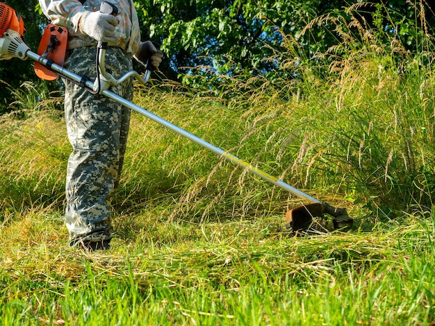 Un hombre vestido con ropa de trabajo usando un cortacésped de gasolina corta hierba larga mantenimiento de la zona rural de verano del territorio