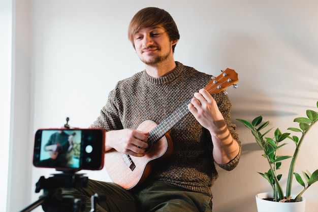 Hombre vestido con un suéter saca el vlog de un juego en un ukelele en casa por teléfono.