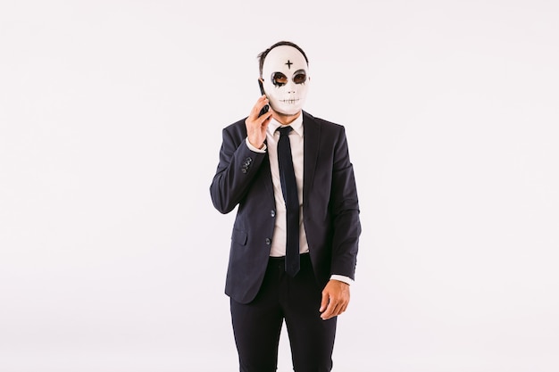 Hombre vestido con traje y corbata, con una cruz de Halloween en su frente máscara asesina, hablando por su teléfono móvil. Celebración de carnaval y halloween