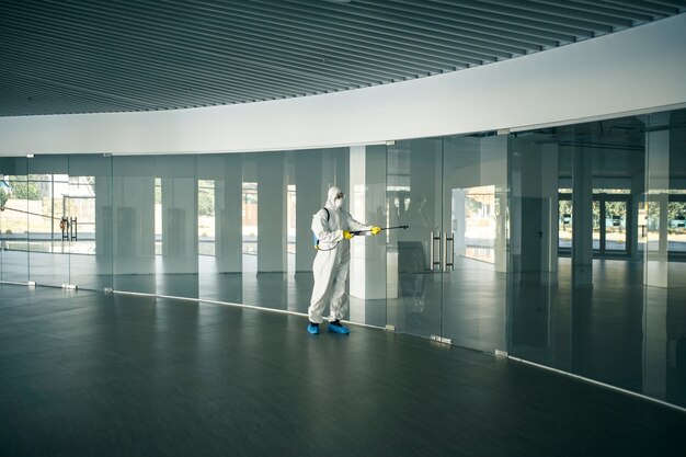 Un hombre vestido con traje de desinfección rocía con desinfectante las manijas de las puertas de vidrio en un centro comercial vacío para evitar la propagación del covid-19. Concienciación sobre la salud, concepto limpio, de defensa.