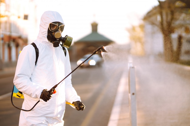 Un hombre vestido con un traje especial de desinfección rocía un esterilizador en el lugar público vacío al amanecer en la ciudad de cuarentena. COVID-19. Concepto de limpieza.