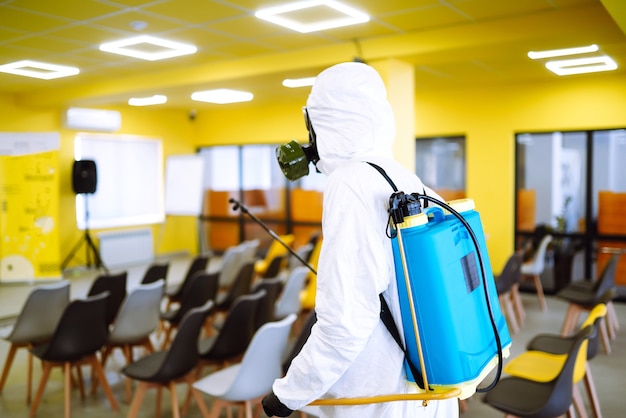 Hombre vestido con traje de protección que desinfecta el salón de actos con productos químicos en aerosol para prevenir el coronavirus.